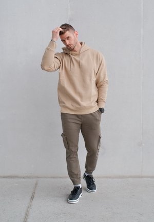 Hombre con sudadera beige con capucha y pantalones cargo caminando en interiores, tocándose el cabello con una mano, frente a un fondo de pared lisa de color gris claro.