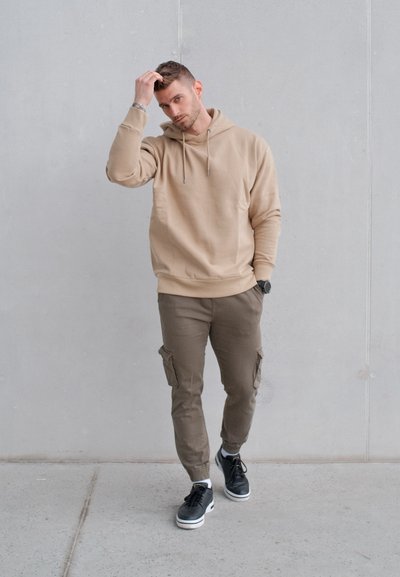 Hombre con sudadera beige con capucha y pantalones cargo caminando en interiores, tocándose el cabello con una mano, frente a un fondo de pared lisa de color gris claro.