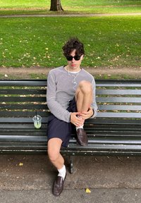 Stripete langermet skjorte, marineblå pinstripe shorts, hvite sokker, brune loafers. Klar kopp med grønn drikke på benk, gressbakgrunn.