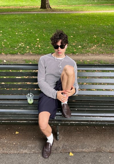 Camisa de rayas de manga larga, pantalones cortos azul marino a rayas, calcetines blancos, mocasines marrones. Vaso transparente con bebida verde sobre un banco, fondo de hierba.