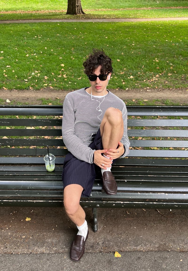 Stribet langærmet skjorte, marineblå stribede shorts, hvide sokker, brune loafers. Klar kop med grøn drik på bænken, græsbaggrund.