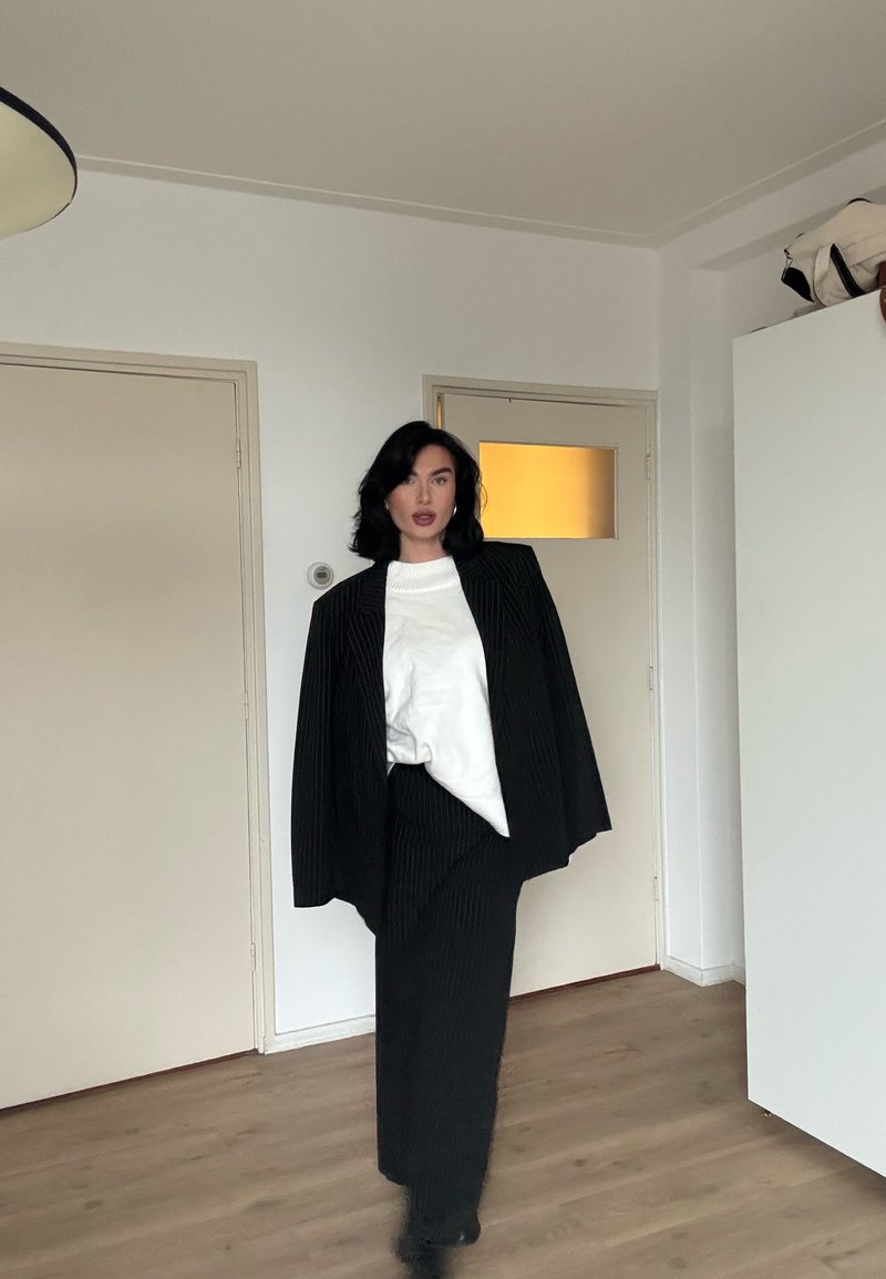 Femme aux cheveux foncés portant un blazer à fines rayures noires et une jupe, avec un haut blanc, debout dans une pièce minimaliste avec un sol en bois et des portes fermées.