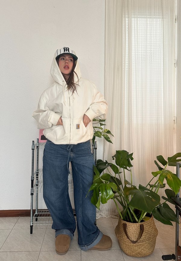 Sweatshirt creme com cordões, bolso frontal; jeans de perna larga azuis; chinelos castanhos; boné com listras pretas e brancas; plantas de interior.