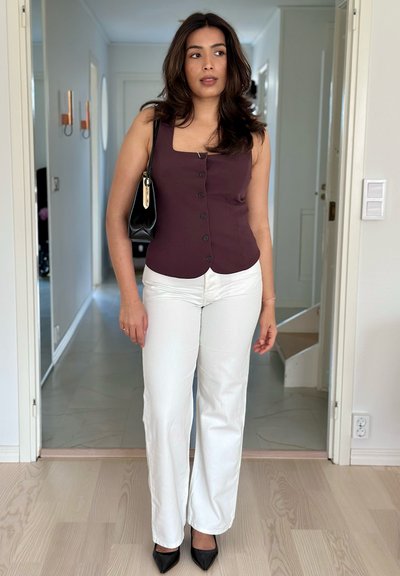 Top burdeos sin mangas con botones, combinado con pantalones blancos de campana. Bolso negro con detalles en dorado, tacones negros de punta. Ambiente interior brillante.