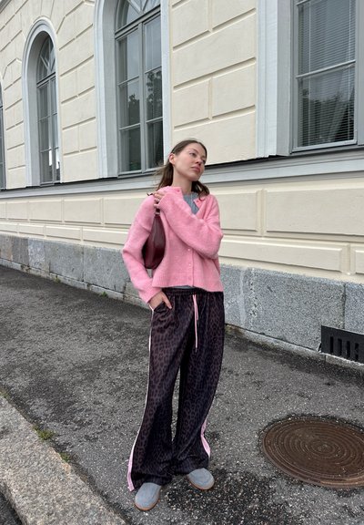 Cardigan de punto rosa, camisa gris, pantalones de leopardo negros con rayas rosas, y zapatos grises sin cordones. De pie sobre una superficie pavimentada.