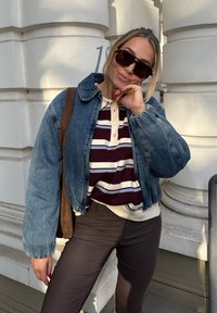 Farkkutakki, jossa on kauluskanta, päällä maroon-raitainen polo ja vaalean beige neule, yhdistettynä tummiin raitapolkkaisiin housuihin ja ruskeaan laukkuun.