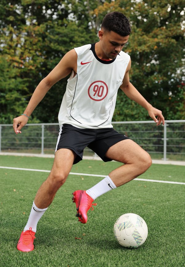 Witte sportieve tanktop met rood logo, zwarte shorts, rode voetbalschoenen en witte sokken, die een patroonvoetbal dribbelt op kunstgras.