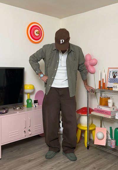 Camisa de cuadros marrón sobre una camiseta blanca, combinada con pantalones marrones holgados y zapatos verdes. Gorra marrón y accesorios plateados.