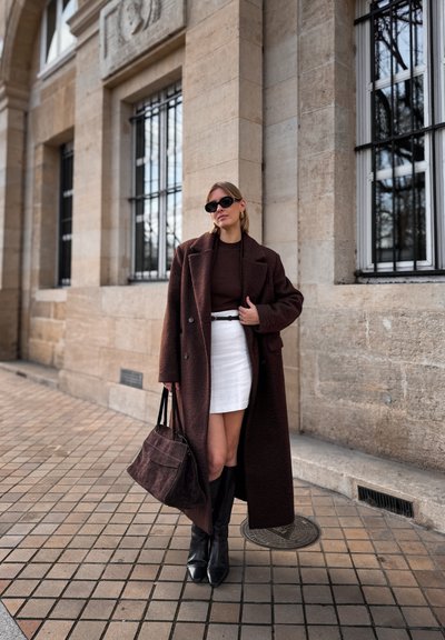 Abrigo de lana marrón sobre un top marrón, minifalda blanca, botas negras hasta la rodilla y un bolso marrón. Las características incluyen texturas suaves y patrones sutiles.