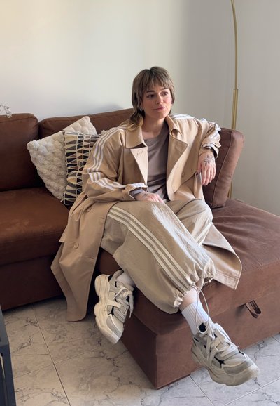 Trinchera beige claro con rayas blancas, combinada con pantalones de tiro ajustable a juego. Zapatillas blancas y un cojín texturizado sobre un sofá marrón.
