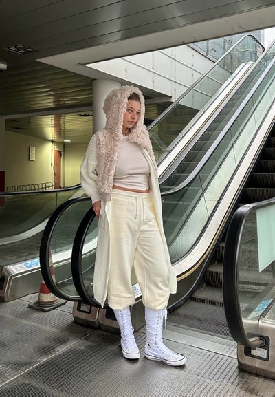 Cárdigan blanco de gran tamaño con capucha de peluche, pantalones claros a juego y zapatillas altas blancas, de pie cerca de una escalera mecánica.