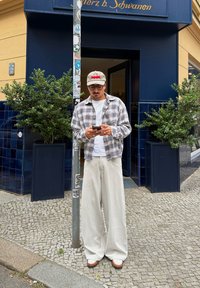 Una persona está de pie al aire libre, vistiendo una camisa de cuadros, una camiseta blanca, pantalones anchos beiges y una gorra gris claro. Están sosteniendo un teléfono inteligente.