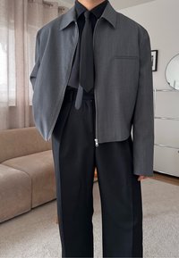 Personne portant une chemise noire et une cravate avec une veste grise courte partiellement zippée et un pantalon noir, debout à l'intérieur près d'un canapé beige.