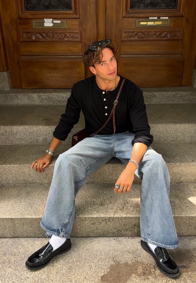 Camisa henley de manga larga negra, pantalones vaqueros de pierna ancha azul claros, mocasines negros y calcetines blancos; sentado en escalones de piedra con puertas de madera al fondo.