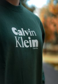 Grønn hettegenser laget av bomull, med en stor hvit "Calvin Klein" logo plassert midt på. Stoffet har en glatt tekstur.