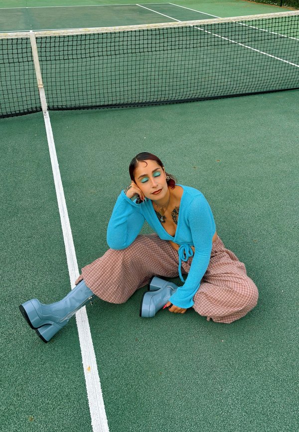 Persona con maglietta blu, pantaloni a fantasia e stivali blu seduta su un campo da tennis vicino alla rete con gli occhi chiusi.