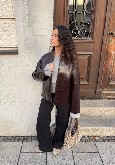 Mujer con cabello largo y ondulado se encuentra en la acera, vestida con una chaqueta de cuero marrón oscuro, un suéter gris, pantalones negros y zapatos beige, sosteniendo un bolso de mano con patrón.