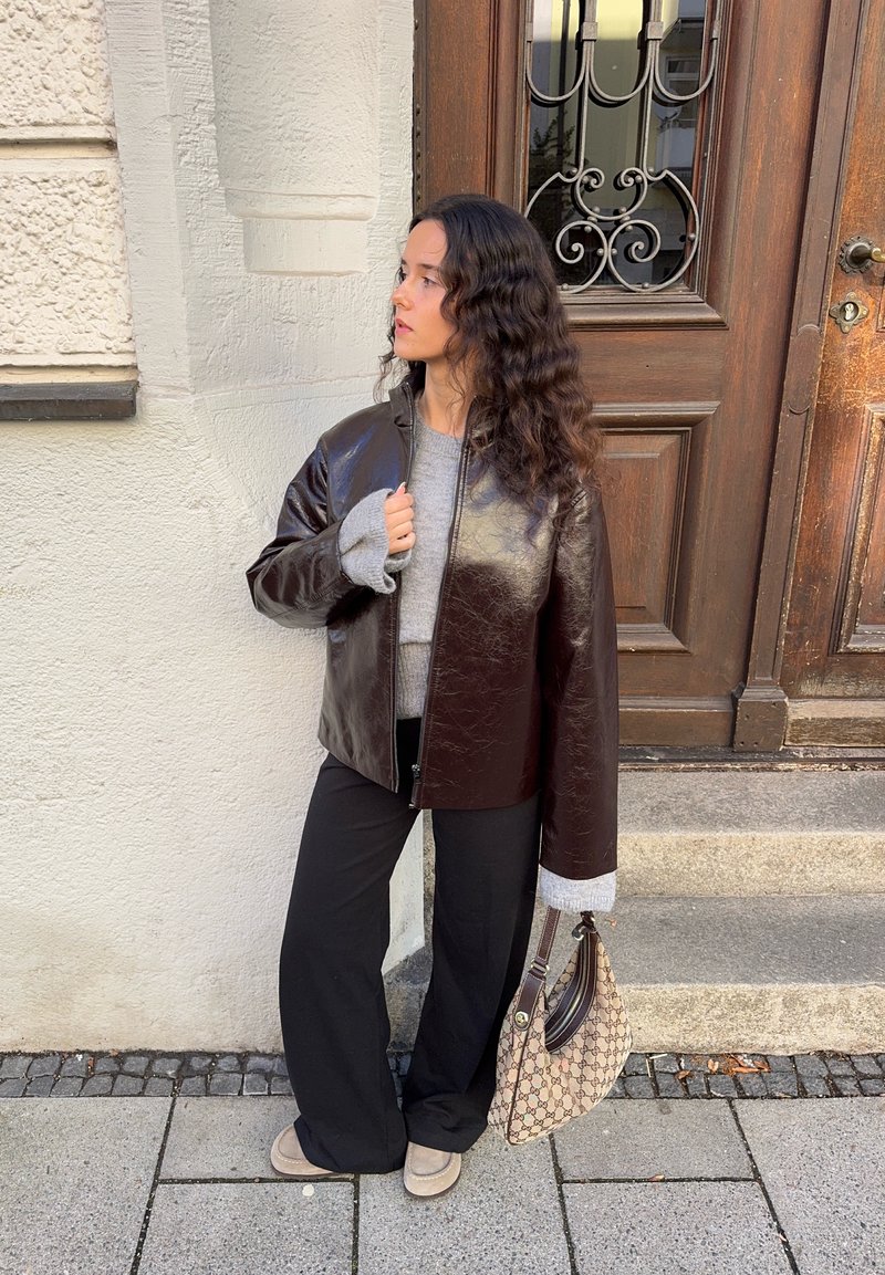 Femme aux longs cheveux ondulés se tient sur le trottoir, vêtue d'une veste en cuir marron foncé, d'un pull gris, d'un pantalon noir et de chaussures beiges, tenant un sac à main à motifs.