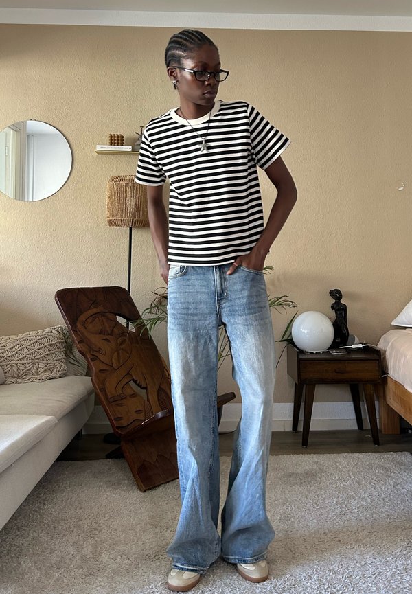 Persona con capelli intrecciati che indossa occhiali, maglietta a righe bianche e nere, jeans a zampa larga, in piedi in un soggiorno con mobili e decorazioni.