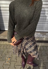 Ciemnozielony, teksturowany sweter dzianinowy zestawiony z kratkowaną spódnicą. Bordowa, błyszcząca mini torebka i wysokie, sięgające do kolan, szpiczaste botki. Wzory obejmują kratkę.