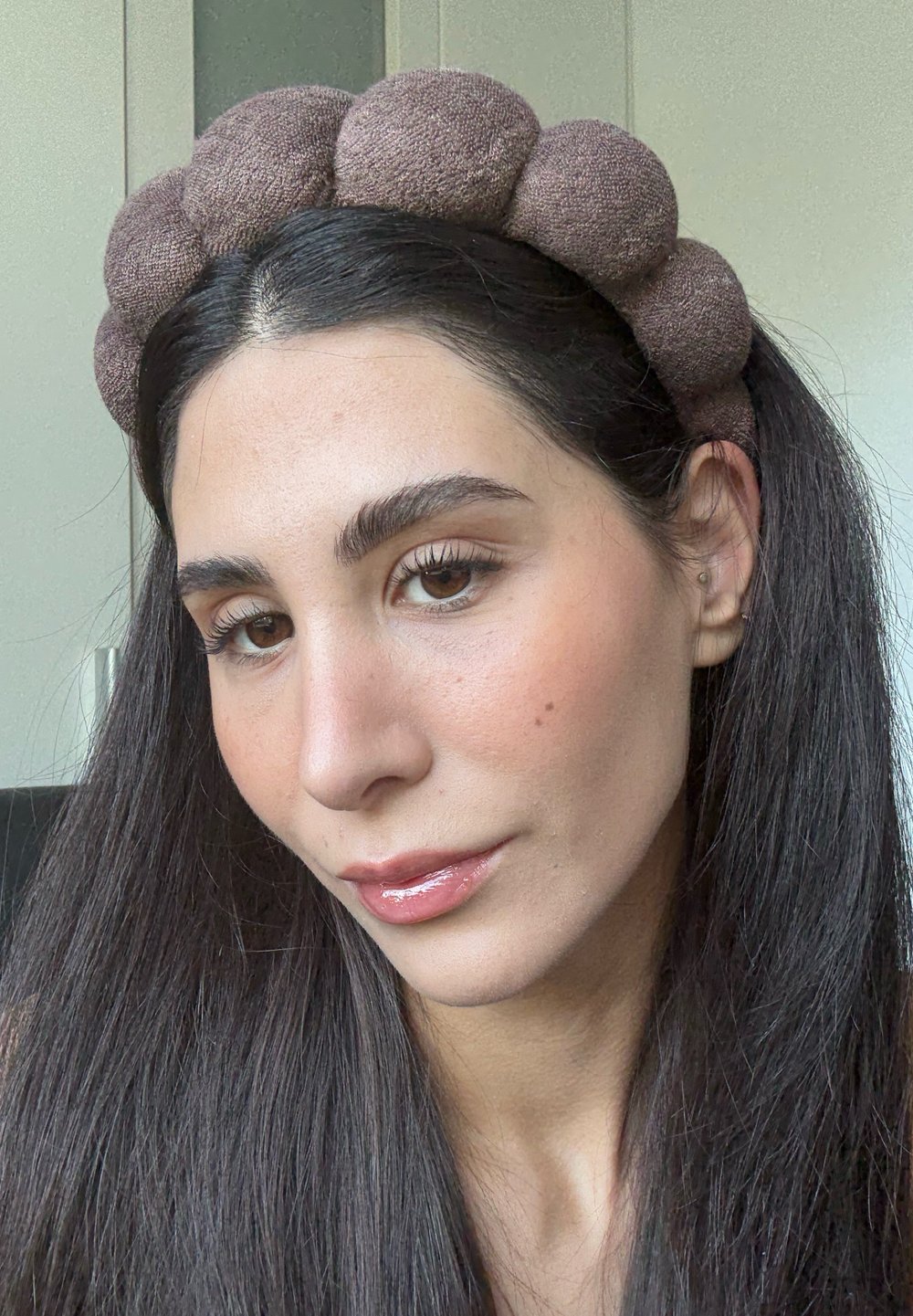 Femme aux longs cheveux bruns portant un bandeau rembourré marron, avec un maquillage naturel et des lèvres brillantes, regardant légèrement vers le bas et souriant doucement.