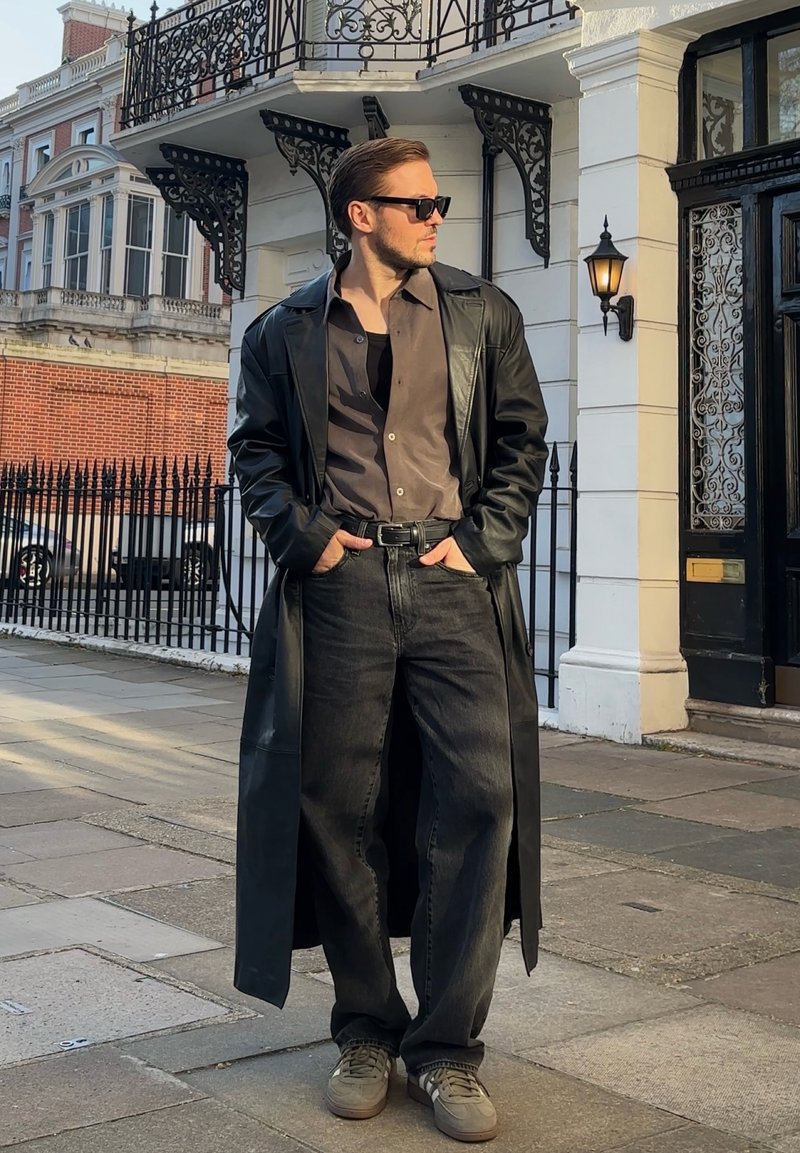 Homme en long manteau en cuir noir, chemise sombre, jean noir ample et baskets, debout sur le trottoir de la ville, les mains dans les poches, regardant sur le côté.