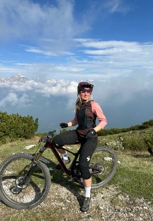 Bicicleta de montaña con un marco marrón, neumáticos negros y un ciclista vestido con una camiseta rosa, equipo de protección negro y un casco morado en un terreno cubierto de hierba.
