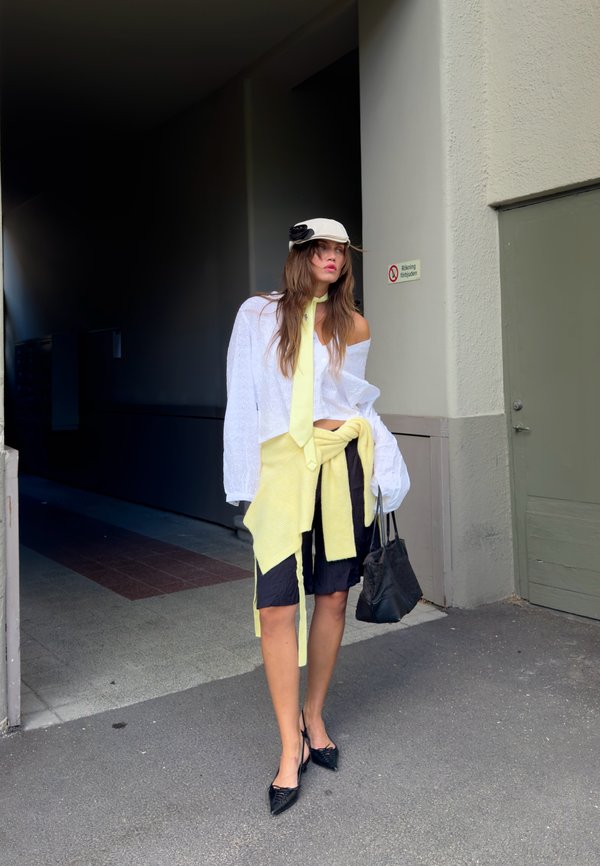 Femme portant un haut blanc et une casquette avec une cravate jaune et un pull autour de la taille, une jupe noire et des talons noirs, debout sur un trottoir près d'un bâtiment.