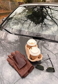 To take-away kaffekopper i en holder, brune skinnhansker med en lommebok oppå, og svarte solbriller på bilens panser som reflekterer trekvister.