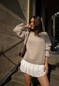 Lys beige chunky sweater med ribbet kant, parret med en hvid lagdelt rynket nederdel. En bordeaux crossbody taske og naturligt lys fremhæver teksturerne.