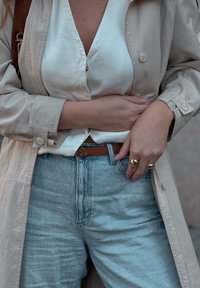 Μπεζ παλτό τρέντσα πάνω από λευ�κή μπλούζα, μπλε τζιν ψηλής μέσης, και καφέ δερμάτινη ζώνη με χρυσό λογότυπο αγκράφα; χρυσά δαχτυλίδια στα δάχτυλα.