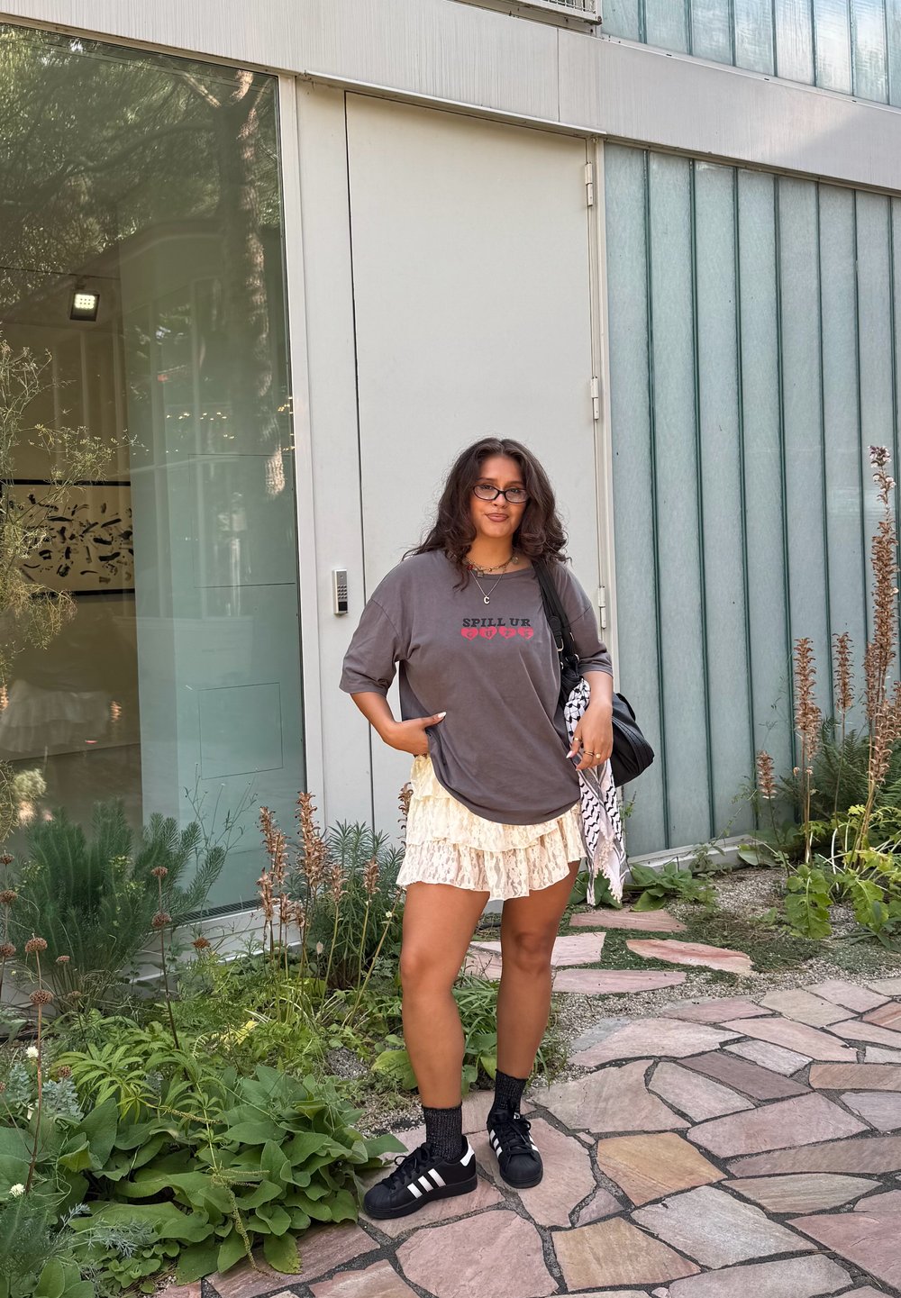 Tricou grafic supradimensionat gri cu textul "SPILL UR", fustă crem cu volane suprapuse, șosete negre și adidași negri. Stă în picioare lângă un perete de sticlă.