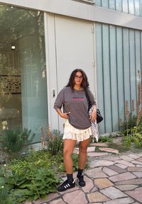 Tricou grafic supradimensionat gri cu textul "SPILL UR", fustă crem cu volane suprapuse, șosete negre și adidași negri. Stă în picioare lângă un perete de sticlă.