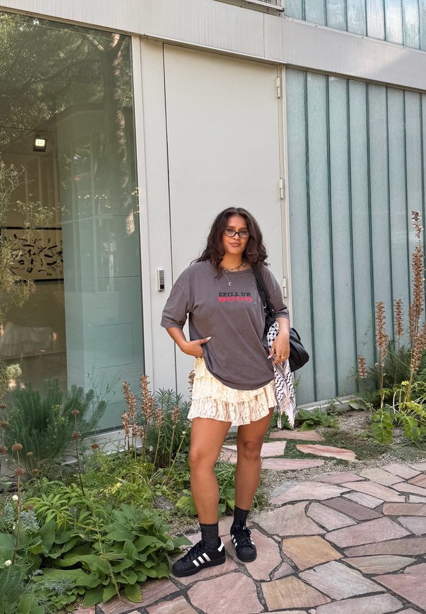 T-shirt oversize gris avec le texte "SPILL UR", jupe crème à volants superposés, chaussettes noires et baskets noires. Debout contre un mur en verre.