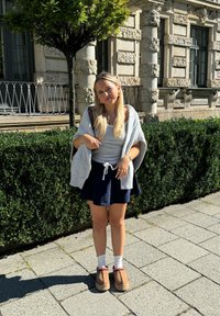 Szary, oversize kardigan noszony na jasnoszarym topie i granatowej spódnicy, białe skarpetki z koronkowym wykończeniem oraz brązowe buty na slip-on z czerwonymi akcentami.