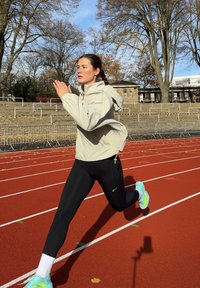 Ljusbeige löparjacka, svarta leggings och turkosa löparskor. Ljusstark löparbana, höstlöv och blå himmel i bakgrunden.