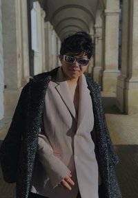 Sacou tailored roz deschis, purtat sub un palton texturat de culoare neagră cu un model herringbone. Ochelari de soare lucioși. Fundal cu un trotuar din piatră arcuit.
