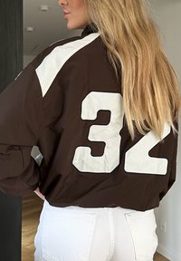 Brun nylonjakke med hvide accenter og nummeret "32" iøjefaldende på ryggen. Har en afslappet pasform og klassisk sporty design.