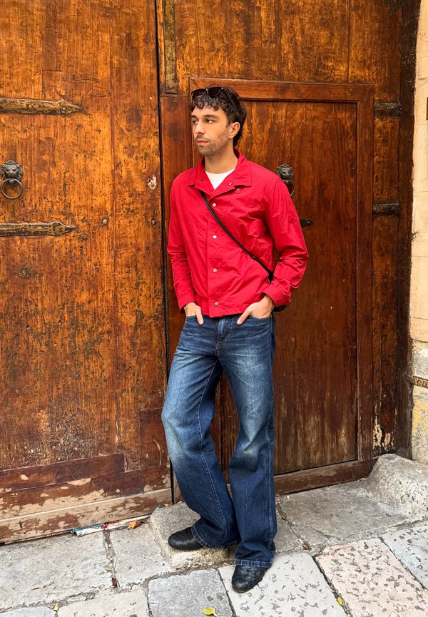 Un homme en veste rouge et en jean se tient les mains dans les poches devant une vieille porte en bois.