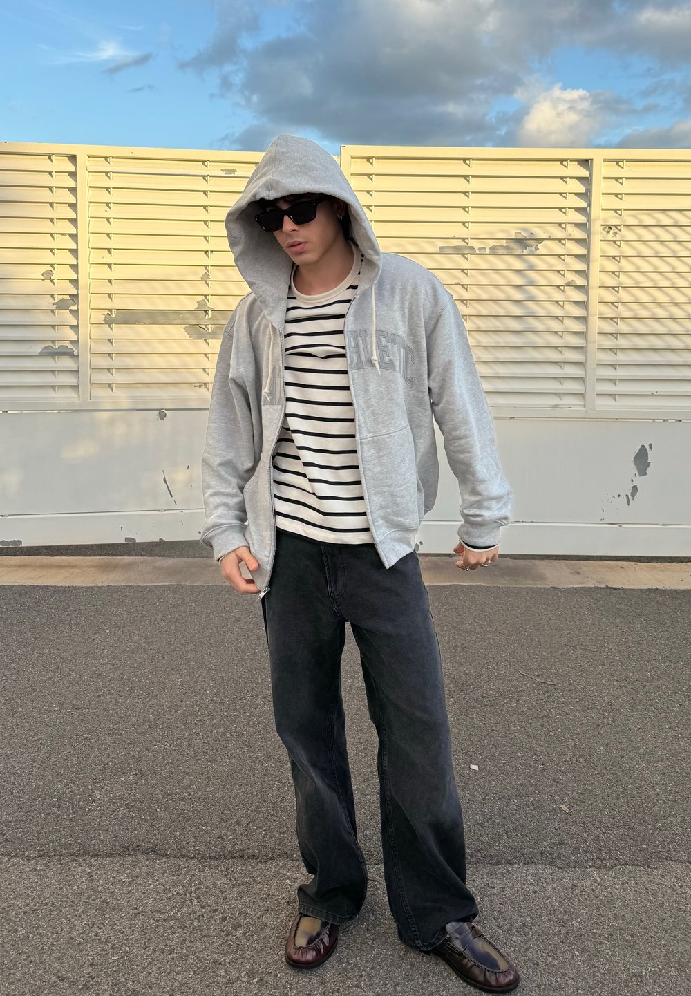 Sweatshirt cinza com fecho, com um bolso frontal, usado sobre uma camisola às riscas, combinado com umas calças largas pretas e sapatos escuros, em pé ao ar livre.
