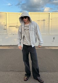 Sweatshirt cinza com fecho, com um bolso frontal, usado sobre uma camisola às riscas, combinado com umas calças largas pretas e sapatos escuros, em pé ao ar livre.
