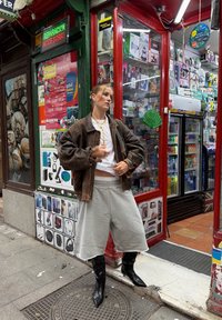 Mulher jovem vestindo uma jaqueta oversized castanha, uma camisa branca, calções largos cinzentos e botas pretas, em pé em frente a uma loja de eletrónicos colorida.