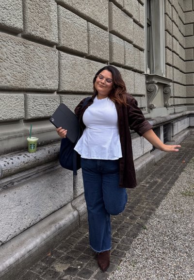 Mujer equilibrándose en una pierna con los ojos cerrados, sosteniendo una tableta, de pie junto a la pared de un edificio de piedra con una bebida de Starbucks cerca.