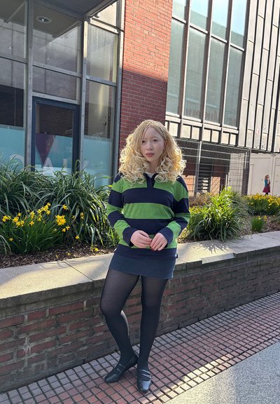 Camisa de rayas de manga larga en verde y azul marino, combinada con una falda de color azul oscuro. Posando al aire libre contra una pared de ladrillo, con plantas y flores cerca.