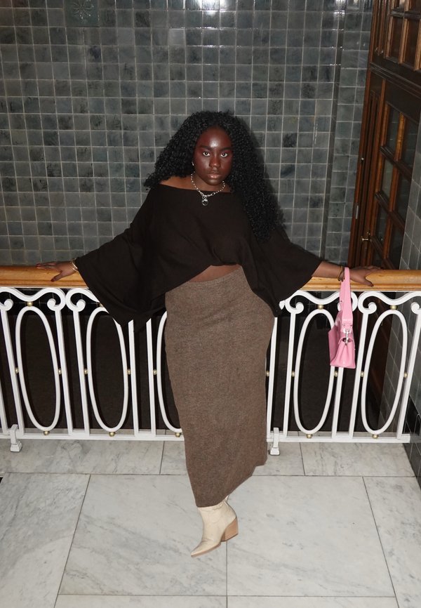 Kvinde med krøllet sort hår læner sig mod hvid gelænder, iført sort bluse med brede ærmer, langt brunt skørt, beige støvler og holder en lyserød håndtaske.