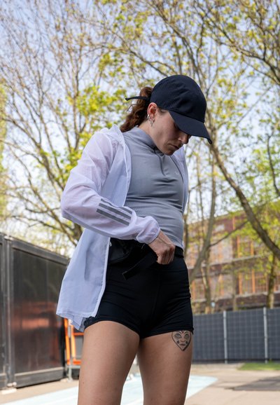 Conjunto atlético: camiseta ajustada de color gris claro, pantalones cortos negros, chaqueta ligera blanca con tres rayas grises. La gorra negra acentúa el look.