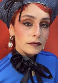Persona con capelli rossi che indossa un cappello blu a righe, grandi orecchini a forma di cuore, una sciarpa floreale scura attorno al collo e una maglietta blu, su uno sfondo rosso.