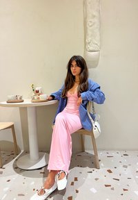 Une personne assise portant une robe rose et une chemise à rayures bleues, avec des chaussures blanches, dans un café minimaliste aux murs clairs et avec un sol en terrazzo.