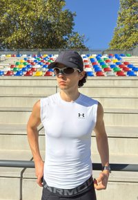 Atletski moški v beli majici brez rokavov, črni kapici in sončnih očalih stoji blizu tribun stadiona s pisanimi vrstami pod jasnim modrim nebom.