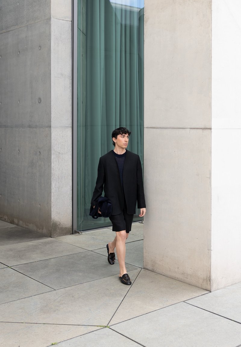 Jeune homme en blazer noir et short marchant sur une place moderne en béton à côté d'un grand bâtiment en verre et béton.